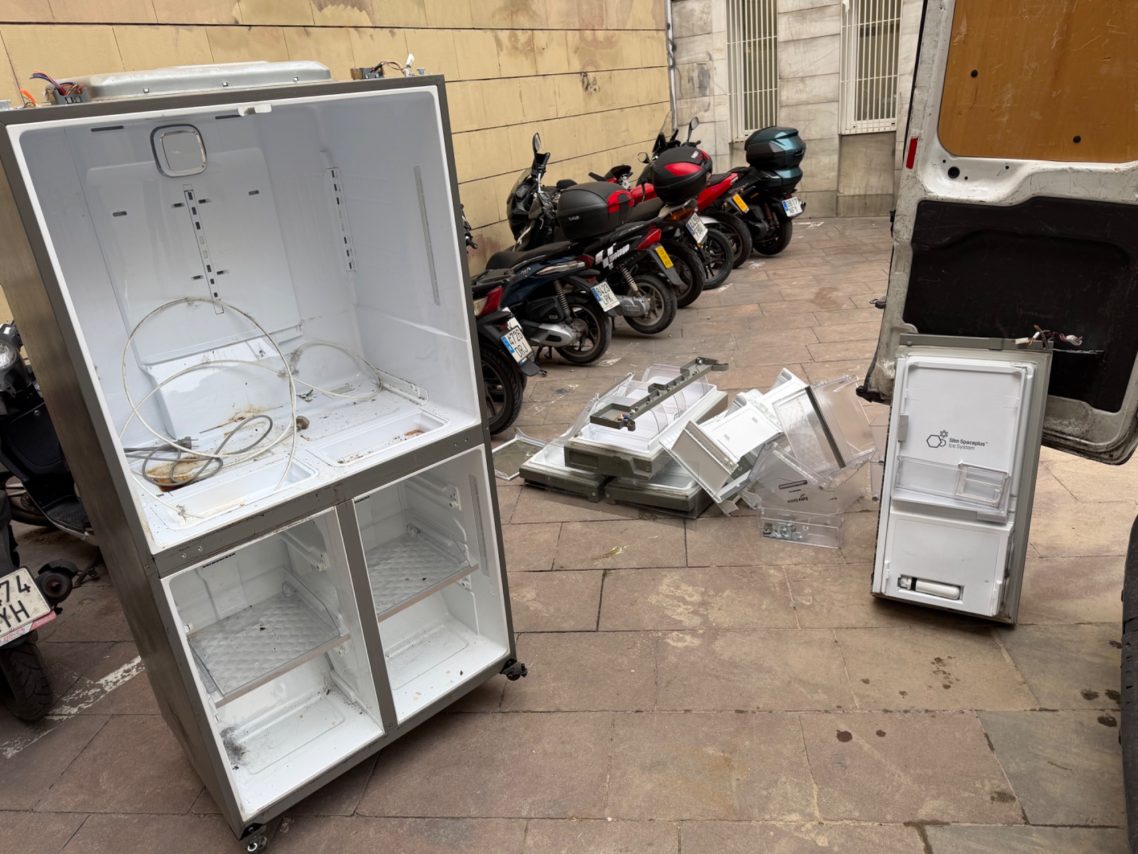 Frigorífico vacío y desarmado en la calle, junto a varios vehículos aparcados.