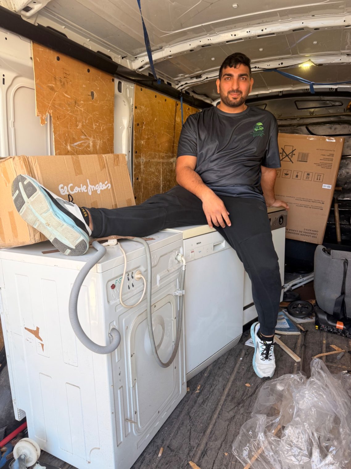 Hombre sentado sobre una lavadora en una furgoneta con cajas en el fondo.