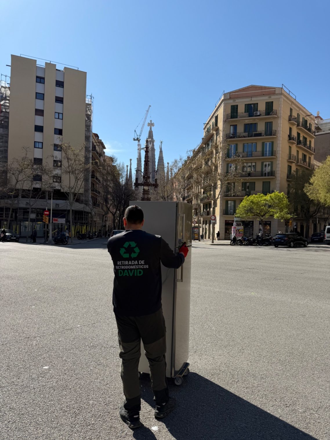 Hombre empujando un carrito por una plaza con edificios y la Sagrada Familia al fondo.